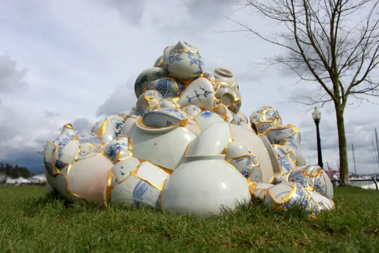 Broken porcelain pots fused together with golden lines in a large, bulbous pile on grass with a winter sky and tree in the background.