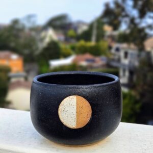 A rice bowl with matte black glaze and a third quarter moon in speckled white over a circle of unglazed speckled buff clay sits on a railing overlooking a blurry hill in the background.