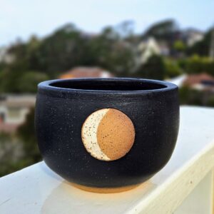 A matte black rice bowl with crescent moon in speckled white over a tan unglazed circle in center of belly sits on a railing with a blurry hillside in the distance.