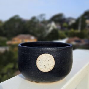 A rice bowl with matte black glaze and a full moon in white over a circle of unglazed speckled buff clay sits on a railing overlooking a blurry hill in the background.