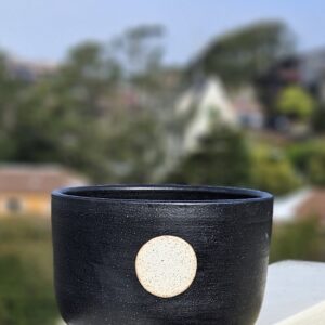 A planter with matte black glaze and a full moon in speckled white over a circle of unglazed speckled buff clay sits on a railing overlooking a blurry hill in the background.