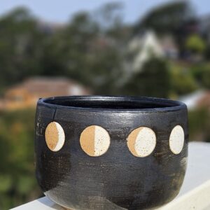 A rice bowl with matte black glaze and four phases of the moon in white over a circle of unglazed speckled buff clay sits on a railing overlooking a blurry hill in the background.