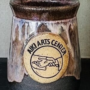 Aiki Arts Center logo shows two hands, one grabbing the wrist of the other. Both wear loose sleeves. Logo is stamped and painted with black ink in a circle of clay that has been applied to a mug with pink, blue and brown glaze over a black clay body.