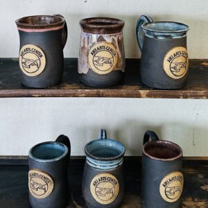 mugs with charcoal claybody, glossy pink or blue glaze inside and around rim, and circular Aiki Arts Center logo in relief in center